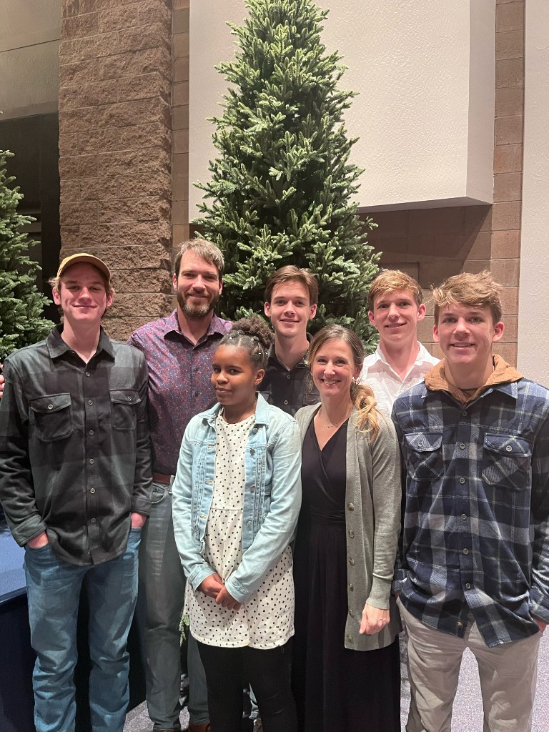 Family of seven posed in front of a tall evergreen indoors