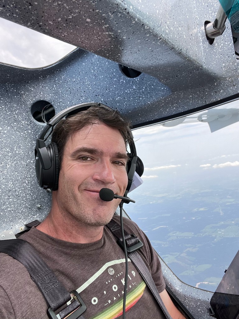 Pilot in the cockpit wearing a headset, fields visible below the wing