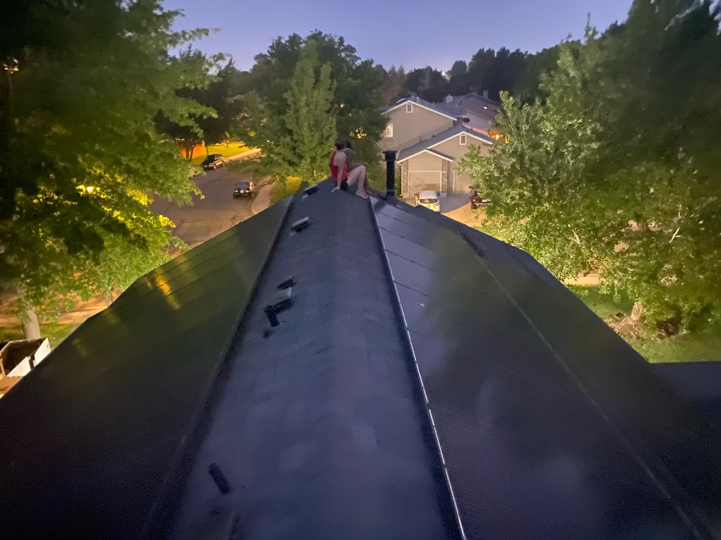 Person on a residential roof ridge at dusk with solar panels and neighborhood lights